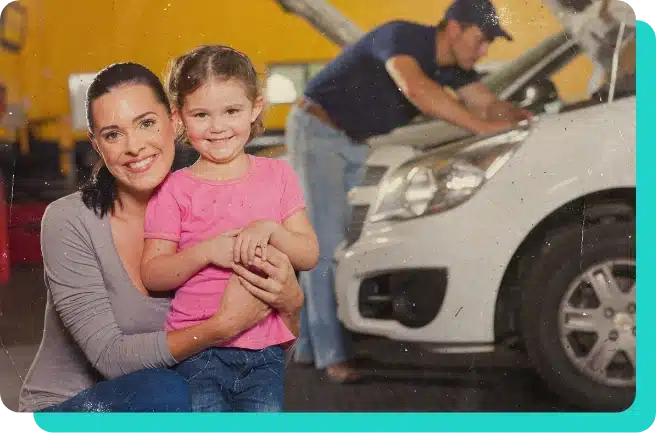 Image of young smiling mother hugging her smiling toddler daughter while a mechanic fixes her white car with hood open in the shop.